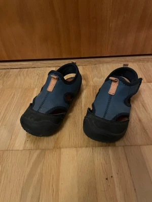 Mörkblå sportiga sandaler - Säljer ett par mörkblå sandaler med svarta detaljer och bruna dragflikar. De har stängd tå, öppna sidor och robust gummisula. Perfekta för en aktiv stil och passar bra till sommarens äventyr.
