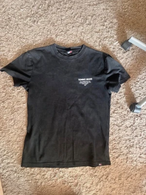 Svart t-shirt från Tommy Jeans - Snygg svart t-shirt från Tommy Jeans med tryck på bröstet och ryggen. Klassisk passform med rund hals och korta ärmar. Perfekt till jeans eller shorts för en enkel och stilren look. Öppen för prisförslag 