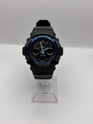Svart G-Shock klocka med blå detalj - Tuff svart G-Shock klocka med blå ring runt urtavlan och digitala samt analoga funktioner. Klockan har robust design, tydliga knappar på sidan och ett brett armband i syntetmaterial. Perfekt för dig som gillar sportig och modern stil.