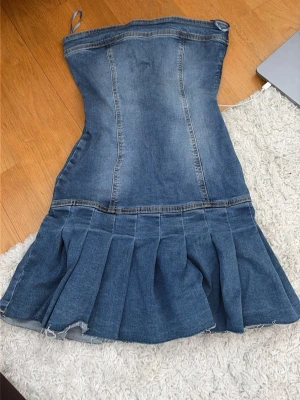 Kort jeansklänning med volang - Trendig axelbandslös jeansklänning i blå denim med figurnära passform upptill och volangig kjoldel. Klänningen har en croppad look och råa kanter nertill för en avslappnad vibe. Perfekt för sommarens alla tillfällen.