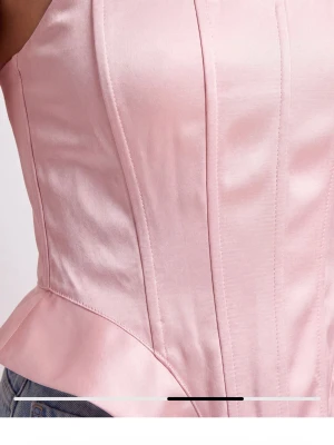 Ljusrosa satin korsett med volang - Supersnygg ljusrosa korsett i glansigt satinmaterial med markerade sömmar och volangdetalj vid midjan. Helt ny och med prislapp på! Figurnära passform och breda axelband ger en modern siluett. Perfekt att styla med jeans för en trendig look.