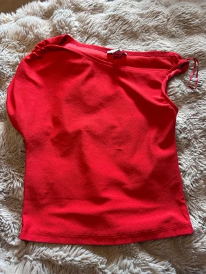 Röd oneshoulder topp - Säljer en snygg röd oneshoulder topp med enkel design. Toppen är ärmlös på ena sidan och har en axelbandslös look på andra sidan. Perfekt för dig som vill sticka ut med en färgstark och trendig topp.