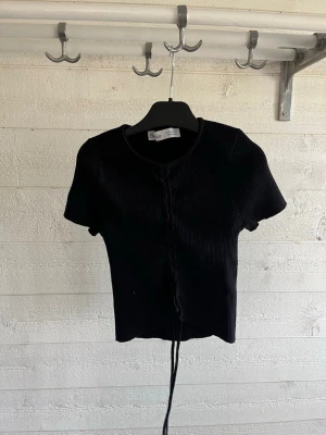 Svart ribbad topp från Bershka - Säljer en svart ribbad topp från Bershka i storlek XS. Toppen har korta ärmar, rund halsringning och snörningsdetalj framtill. Perfekt för dig som gillar en enkel men ändå trendig stil.