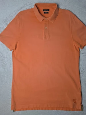 Massimo Dutti Casual Fit piké orange storlek L - Orange pikétröja från Massimo Dutti i Casual Fit-passform. Klassisk enfärgad modell i pikéstickad bomull med diskret ton-i-ton-broderad logotyp nedtill framtill.  Storlek L Casual Fit  Mycket fint skick.  Mått Bröstbredd: ca 52 cm Plagglängd: ca 74 cm Axelbredd: ca 45 cm  Material 100% bomull  Klassisk pikékrage, kort ärm, två knappar framtill och ribbade avslut vid krage och ärmar.  Varan är begagnad och säljs i befintligt skick, se bilder för detaljer. 