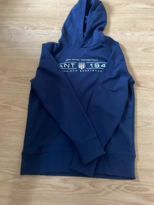 Mörkblå hoodie från GANT - Snygg mörkblå hoodie från GANT med tryck på bröstet där det står 'GANT 1949 American Sportswear'. Klassisk huva och långa ärmar. Perfekt för en avslappnad och sportig stil. Tillverkad i mjukt material för extra komfort.