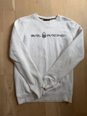 Vit sweatshirt från Sail Racing - Vit sweatshirt från Sail Racing med svart logotyp och text på bröstet. Tröjan har rund halsringning, långa ärmar och ribbade muddar vid ärmslut och nederkant. Perfekt för en avslappnad och sportig stil.