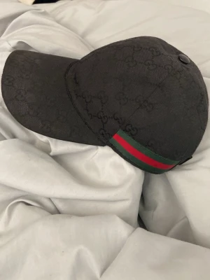 Svart keps med GG-mönster och röd/grön detalj - Snygg svart keps med diskret GG-mönster över hela ytan och en klassisk röd/grön rand på sidan. Kepsen har böjd skärm och justerbar rem baktill. Perfekt för dig som vill ha en trendig accessoar med exklusiv känsla.