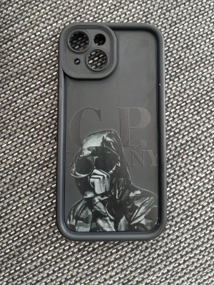 iPhone 15 C.P. Company mobilskal  - Snyggt svart mobilskal med tryck av en person i huva och glasögon samt texten C.P. Company. Skalet har en matt finish och är gjort i tålig plast. Passar perfekt för dig som vill ha en unik och streetinspirerad look på mobilen.