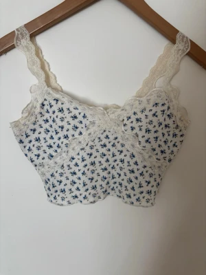 Blommig spetsdetaljerad topp - Söt topp i vitt med små blå blommor och breda spetskanter. Axelbanden är också i spets och tyget är ribbat🤍