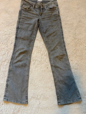 Grå bootcut jeans med låg midja - Säljer ett par grå bootcut jeans med låg midja och klassisk femficksmodell. Jeansen har utsvängda ben och är tillverkade i ett mjukt denimtyg som ger en avslappnad look. Perfekta för dig som gillar retrovibbar och vill ha ett par bekväma jeans till vardags.