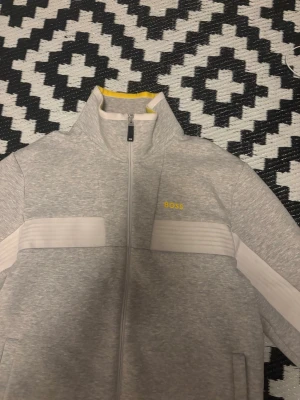 Grå ziptröja från BOSS - Snygg grå ziptröja från BOSS med vita detaljer längs ärmar och bröst. Tröjan har hög krage, dragkedja framtill och gul BOSS-logga på bröstet. Perfekt för en sportig och avslappnad stil.