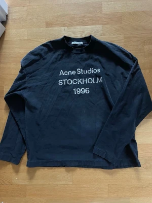 Svart långärmad tröja Acne Studios - Svart långärmad tröja från Acne Studios med vit text 'Acne Studios STOCKHOLM 1996' på bröstet. Tröjan har en avslappnad passform, rund halsringning och är tillverkad i mjuk bomull. Perfekt för en stilren och enkel look. svart/gråmelerad, tvättar såklart innan jag skickar! 