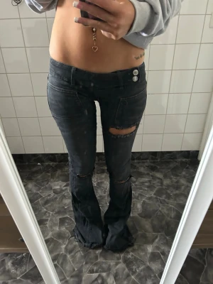 Svarta slitna bootcut jeans - Säljer ett par svarta bootcut jeans med slitningar och hål vid knäna. Jeansen har låg midja, två knappar i linningen. Perfekta för dig som gillar en edgy och trendig stil.