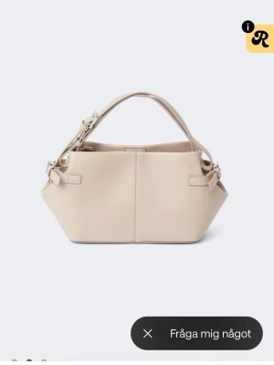 Beige handväska från gina - Stilren beige handväska i slätt konstläder med två handtag och snygga spänndetaljer i silver på sidorna. Väskan har en modern, kantig form och minimalistisk design som passar till många olika looks. Perfekt för dig som gillar enkel men trendig stil.
