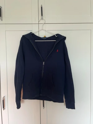 Mörkblå zip hoodie från Polo Ralph Lauren - Mörkblå hoodie med dragkedja från Polo Ralph Lauren. Klassisk design med huva, två fickor framtill och den ikoniska röda loggan broderad på bröstet. Perfekt för en chill och stilren look. Mjuk bomullskänsla och ribbade muddar.