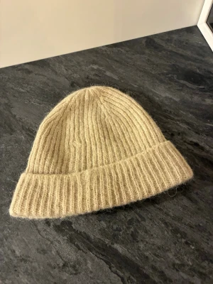 Beige ribbstickad mössa i ull - en Mysig beige mössa i ribbstickat i mörket carin wester materialet är  med uppvikt kant. Perfekt för kalla dagar och ger en avslappnad, trendig look. Tillverkad i mjukt garn som känns skönt mot huden.