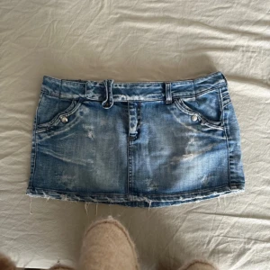 Blå jeanskjol med slitningar och bälte - Säljer en kort blå jeanskjol med slitna detaljer och rå kant. Kjolen har fickor både fram och bak samt ett matchande jeansbälte i midjan. 