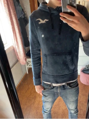 Blå hoodie från Hollister med tryck - Modellen 183 blå hoodie från Hollister med stor logga på bröstet och texten 'HOLLISTER' längs ena ärmen. Hoodien har huva med dragsko och en klassisk magficka. Tillverkad i mjukt material som är skönt att ha på sig. Perfekt för en avslappnad och trendig look.