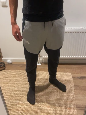 Nike svart-grå mjukisbyxor - Nike mjukisbyxor i svart och ljusgrått med dragsko i midjan och ribbade muddar vid bensluten. Byxorna har en sportig look med kontrastpaneler och Nike-logga på vänster sida. Perfekta för chill eller träning.