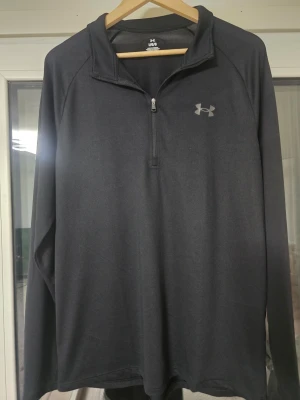 Mörkgrå träningströja från Under Armour - Snygg mörkgrå långärmad träningströja från Under Armour med halv dragkedja framtill och diskret logga på bröstet. Tillverkad i ett lätt och funktionellt material som passar perfekt till träning eller löpning. Enkel och stilren design.