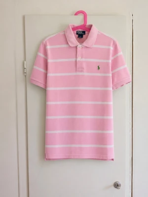 🌸 Rosa randig Polo Ralph Lauren piké 🌸 - Superfin klassisk piké från Polo Ralph Lauren i rosa med vita ränder och den ikoniska gröna broderade loggan på bröstet. Storlek: M (10–12 år) Material: 100 % bomull Färg: Rosa/vit Skick: Mycket fint skick men har en fläck! Perfekt till sommaren och supersnygg för en preppy/old money-look 💗 Hör gärna av dig vid frågor eller om du vill ha mått!