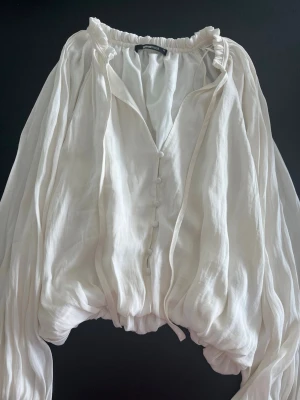 Vit voluminös blus med knappar - Säljer en vit, luftig blus med voluminösa långärmar och knappar framtill. Blusen har en rynkad resår i nederkant och vid halsen, samt ett skirt, lätt material som ger en romantisk känsla. Perfekt för dig som gillar en bohemisk och trendig stil.
