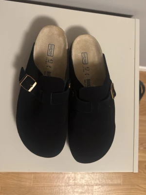 Svarta mocka tofflor med spänne - Säljer ett par svarta tofflor i mocka med spännen. Klassisk slip-in modell med ergonomisk fotbädd och mönstrad sula för bra grepp. Perfekta för chill dagar hemma eller när du vill ha det bekvämt.