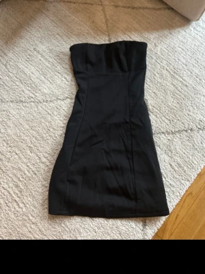 Svart axelbandslös kort klänning - Säljer en svart, axelbandslös kort klänning med figurnära passform. Klänningen har en enkel och stilren design utan synliga detaljer eller mönster, perfekt för dig som gillar minimalistisk stil.