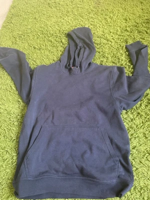 Mörkblå hoodie med huva - En enkel och stilren mörkblå hoodie med stor känguruficka framtill och justerbar huva med dragsko. Hoodien har långa ärmar och ribbade muddar vid ärmslut och nederkant. Perfekt för en avslappnad och bekväm look.