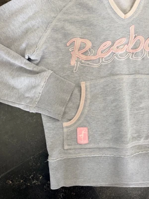 Grå vintage hoodie från Reebok med rosa detaljer - Snygg grå hoodie från Reebok med rosa broderad logga på bröstet och rosa detaljer vid fickor och huva. Klassisk känguruficka framtill och ribbade muddar vid ärmslut och nederkant, tunnare hoodie i vintagemodell. M i storleken på lappen men snarare en 34/36/XS