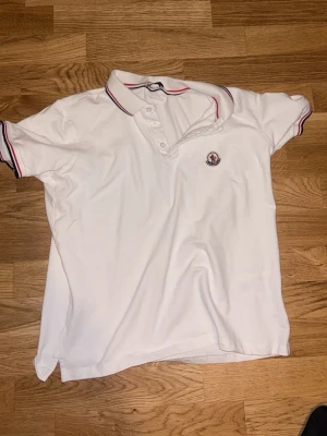 Vit Moncler pikétröja - Klassisk vit pikétröja från Moncler med korta ärmar och krage. Tröjan har röda och marinblå ränder på krage och ärmslut samt Moncler-logga på bröstet. Perfekt till en avslappnad men stilren look.