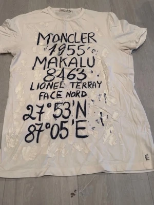 Vit Moncler t-shirt med tryck - Vit t-shirt från Moncler med svart handskriftsinspirerat tryck framtill med text och koordinater. Klassisk rund halsringning och korta ärmar. Perfekt för dig som gillar statement-plagg och streetstyle. Köpt på semester i Italien 