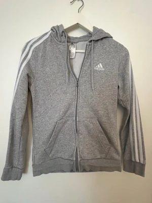Grå zip hoodie från Adidas - Snygg basic grå hoodie från Adidas med dragkedja, huva och vita klassiska ränder längs ärmarna. Adidas-logga på bröstet och två fickor framtill. Tillverkad i mjukt sweatshirtmaterial.