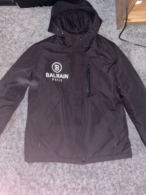 Svart tjockjacka från Balmain Paris - Snygg svart  tjockjacka från Balmain Paris med vit logga på bröstet. Jackan har huva, dragkedja framtill och en bröstficka med dragkedja. Perfekt för blåsiga dagar och streetwear-stil.