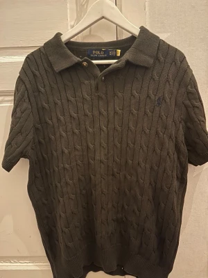 Mörkgrå kabelstickad polotopp - Kabelstickad mörkgrå polotopp från Polo Ralph Lauren med korta ärmar och klassisk krage. Tröjan har knappdetalj vid halsen och diskret broderad logga på bröstet. Perfekt för en stilren och avslappnad look.Priset går att diskutera vid snabb affär, finn de frågor så är de bara o skriv 😊