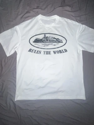 Street wear T shirts 'Rules The World' - Vit t-shirt i bomull med svart tryck framtill som föreställer ett fartyg och texten 'RULES THE WORLD'. Klassisk passform och rund halsringning. Perfekt för dig som gillar statement-plagg och enkel stil.