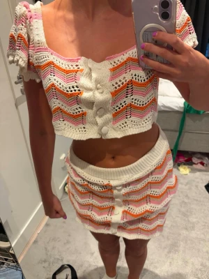 Stickat set i vitt, rosa och orange - Säljer ett superfint tvådelat set med virkad topp och kjol i vitt med vågmönster i rosa, orange och lila. Toppen har korta puffärmar och stora dekorativa knappar framtill. Kjolen är högmidjad och matchar toppen perfekt. Perfekt för sommarens alla tillfällen! En knapp har fallit av på toppen. 