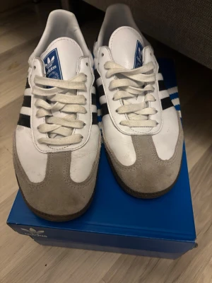 Adidas Samba vita och svarta sneakers - Klassiska Adidas Samba sneakers i vitt läder med grå mockadetaljer och svarta ränder på sidorna. Skorna har vit plös med blå Adidas-logga och svart yttersula. Tidlös design som funkar till det mesta och ger en sportig vibe.
