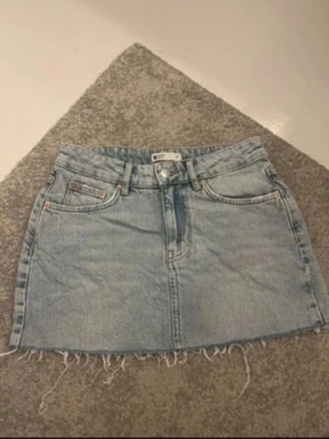 Ljusblå jeanskjol med fransig kant - Säljer en ljusblå jeanskjol i klassisk modell med fem fickor och fransig, rå kant nedtill. Kjolen har bälteshällor och dragkedja framtill. Perfekt till sommaren och enkel att styla med olika toppar.
