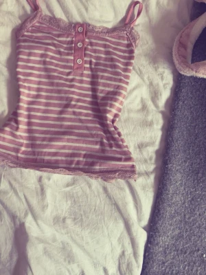 Rosa randigt  linne  med spets - Sött (pyjamaslinne) i rosa och vitt med smala axelband och spetsdetaljer vid halsringning och nederkant. Linnet har tre knappar framtill och är gjort i mjukt, stretchigt material. Perfekt för mysiga kvällar hemma.