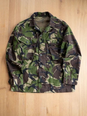 Army Jacket, size M - Snygg overshirt i klassiskt camouflage-mönster med gröna, bruna och beige toner. Skjortan har långa ärmar, krage och knäppning framtill samt två bröstfickor med knappar. Tillverkad i bomull för en skön och slitstark känsla.