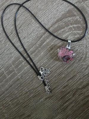 Halsband med rosa blomberlock - Snyggt halsband med svart snöre och en genomskinlig berlock i form av ett hjärta. Berlocken har ett rosa blommotiv inuti som ger en unik och konstnärlig känsla. Justerbart spänne i silverfärg. Perfekt accessoar för dig som gillar naturinspirerade smycken.