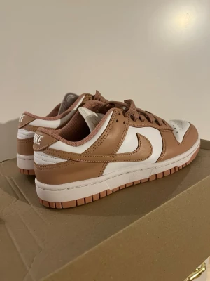 Nike Dunk Low vit/brun sneaker - Nike Dunk Low sneakers i vitt och ljusbrunt läder. Klassisk låg siluett med perforerad tåbox, vita paneler och bruna detaljer på Swoosh, snörning och yttersula. Perfekt för dig som gillar stilrena och trendiga sneakers. Går att diskutera priset vid snabb affär.
