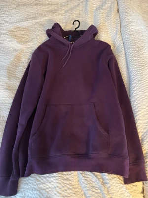 Lila hoodie med huva och magficka - En enkel och stilren lila hoodie med huva och dragsko. Hoodien har en stor magficka framtill och ribbade muddar vid ärmslut och nederkant. Perfekt för dig som gillar en avslappnad och bekväm look. Materialet känns mjukt och skönt mot huden.