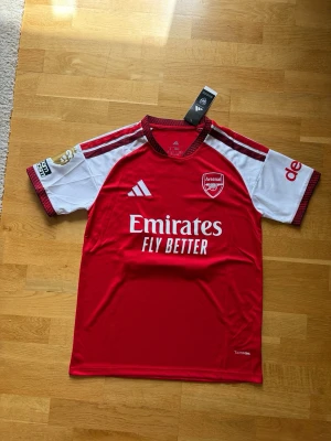 Arsenal matchtröja röd/vit Adidas - Snygg Arsenal matchtröja i rött och vitt från Adidas med klubbmärke och sponsortryck framtill. Tröjan har korta ärmar, rund hals och tryck med namnet Gyökeres och nummer 14 på ryggen. Perfekt för fotbollsfans som vill visa sitt stöd på läktaren eller planen.