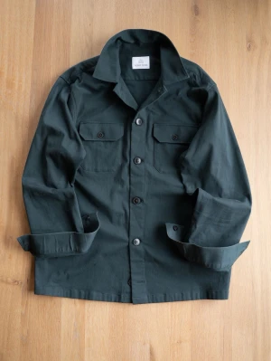 Mörkgrön overshirt från The Shirt Factory - Stilren mörkgrön overshirt från The Shirt Factory med klassisk krage, knäppning framtill och två bröstfickor med lock och knapp. Tillverkad i ett kraftigare bomullstyg som ger en schysst och avslappnad look. Perfekt att bära öppen eller stängd.