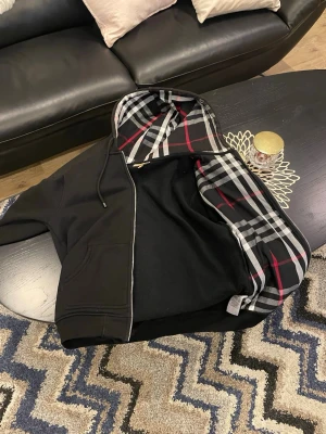 Svart hoodie med rutigt foder - Säljer en svart hoodie burberry dragkedja och stor känguruficka framtill. Insidan av huvan har ett rutigt mönster i svart, vitt och rött som ger en snygg kontrast. Perfekt för dig som vill ha en stilren men ändå unik hoodie.