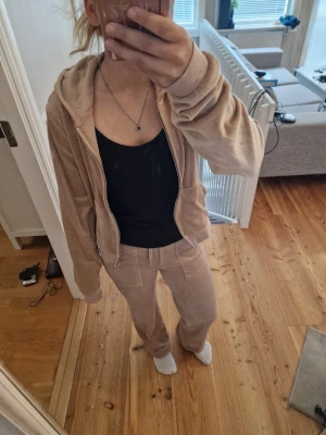Beige Juicy couture set  - Mysigt Juicy couture set i beige velour med zip-hoodie och matchande byxor. Hoodien har dragkedja, fickor och huva, medan byxorna har resår i midjan med snörning och fickor fram. Perfekt för chill dagar hemma. Byxorna är upplagda och personen på bilden är 155cm lång. 