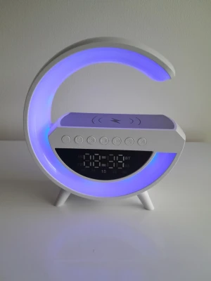 Ny BT 2301 G-formad LED trådlös laddare och Bluetooth-högtalare med FM-radio - Denna helt nya och multifunktionella BT 2301 LED-högtalare med trådlös laddning har en modern och stilren G-formad design som passar perfekt i sovrummet, på kontoret eller i arbetsrummet. Den är fullpackad med mångsidiga funktioner och fungerar som en allt-i-ett-lösning för stämningsbelysning, trådlös laddning och bärbart ljud. ​Trådlös laddningsplatta: Den platta ovansidan har en inbyggd trådlös laddningsplatta (markerad med en blixtsymbol) för att snabbt och smidigt ladda din smartphone. ​Dyna