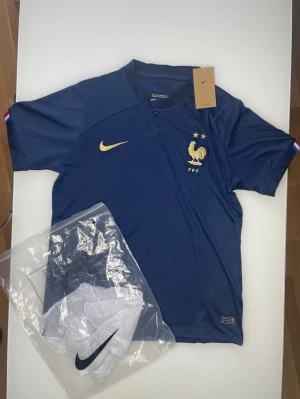 Frankrike Nike matchtröja blå - Snygg blå matchtröja från Nike med Frankrikes landslagsemblem och två stjärnor på bröstet. Guldiga detaljer och korta ärmar. Tillverkad i lätt och ventilerande material, perfekt för fotboll eller träning.